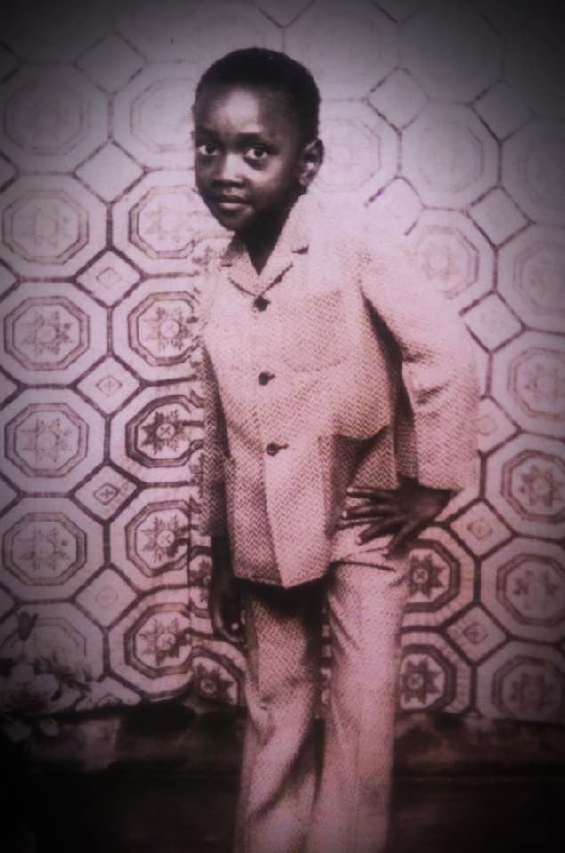 Photo d'archive : Mike Olembo à 5 ans, en costume, posant devant un papier peint